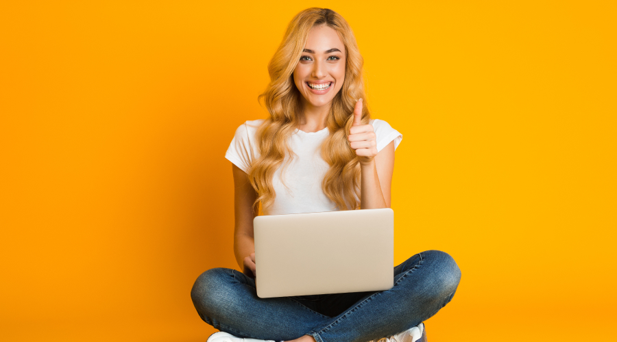 Woman smiling with thumbs up sitting with a laptop against a yellow background