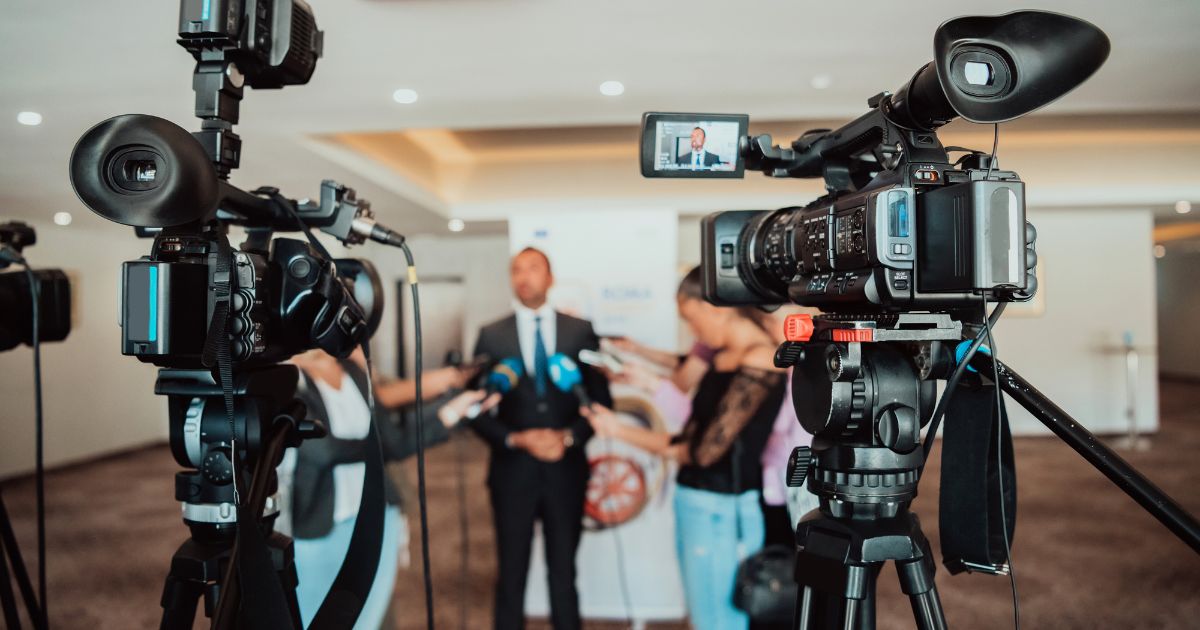 Video cameras filming a corporate press conference with speakers and media crew