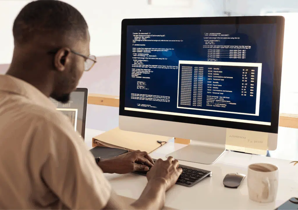 Man sitting at a desk working on a computer screen filled with lines of code in a modern office.