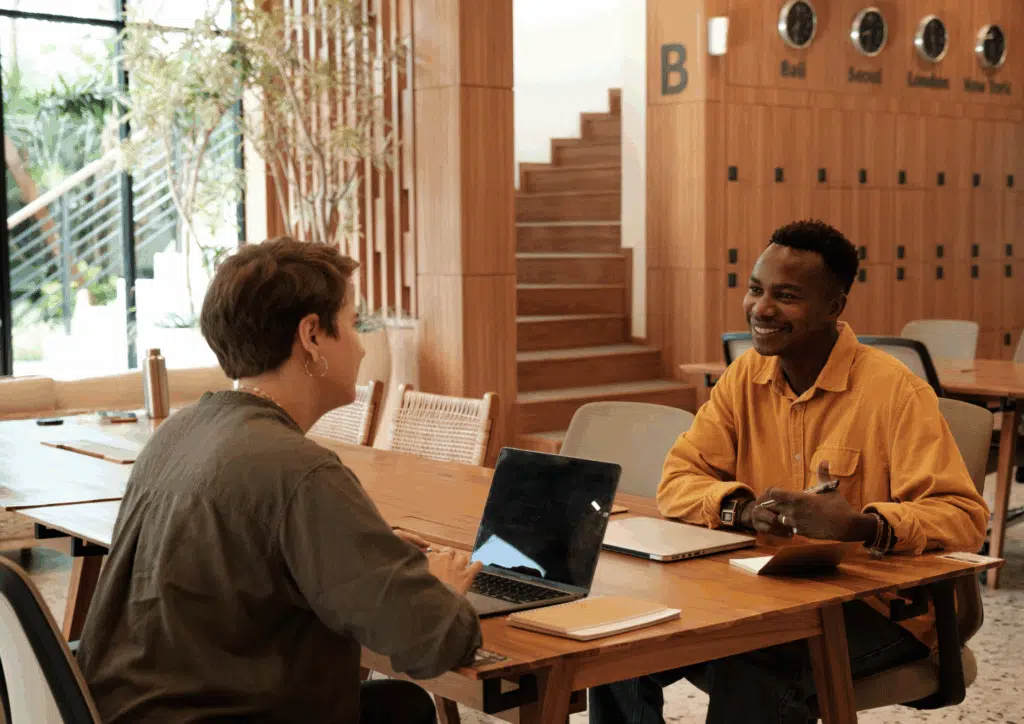 A smiling young man in a mustard shirt sits across from a woman interviewer with a laptop, engaged in a conversation in a bright, wooden office space with natural light and greenery.