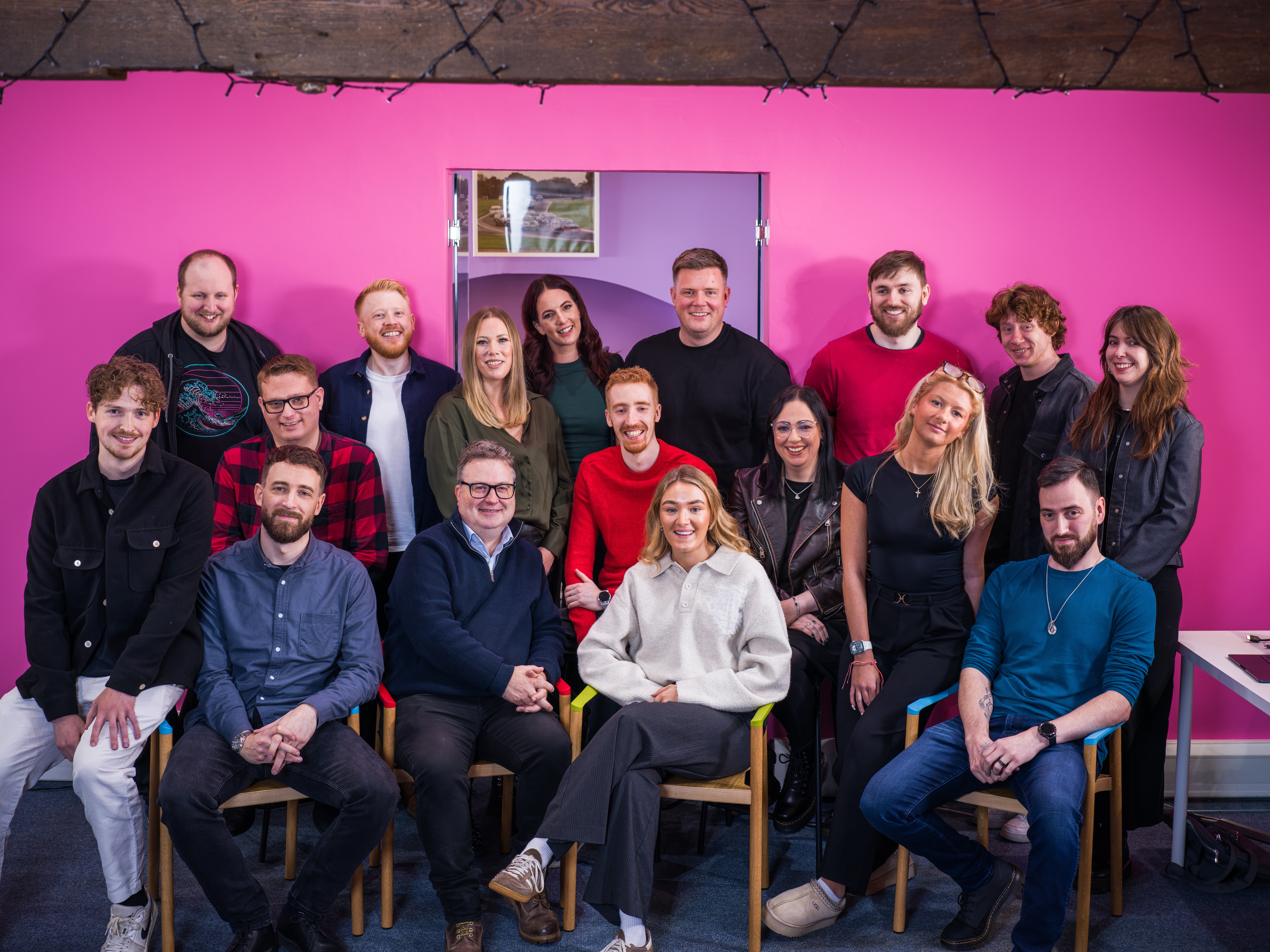 A diverse group of people sitting together in front of a bright pink wall, smiling and enjoying each other's company.