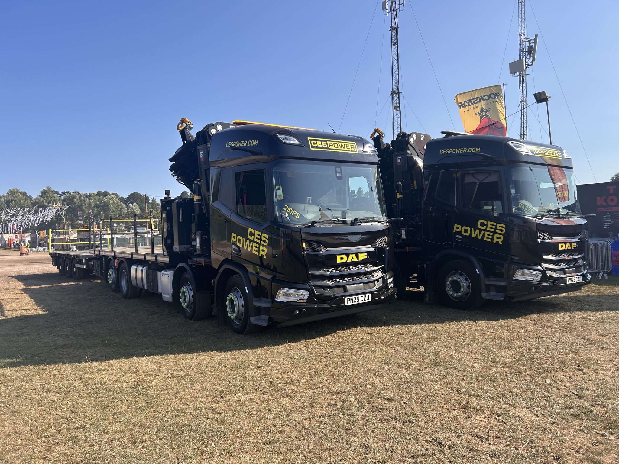 Two black CES Power flatbed trucks parked side by side at an outdoor event site, with lifting cranes mounted behind the cabs and festival infrastructure visible in the background.