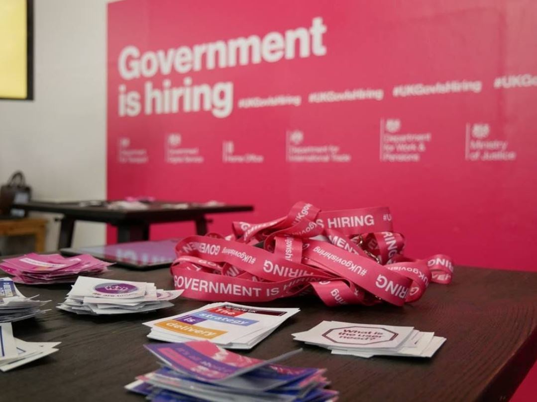 The focus of the image are some lanyards and sticker on a table. They're in front of a pink billboard that reads 'Government is hiring'