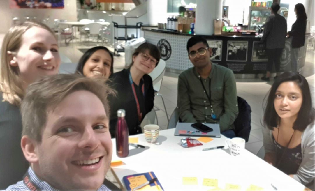 A group of 5 DfE product managers smiling at the camera while sitting down at a table during their product crit meeting
