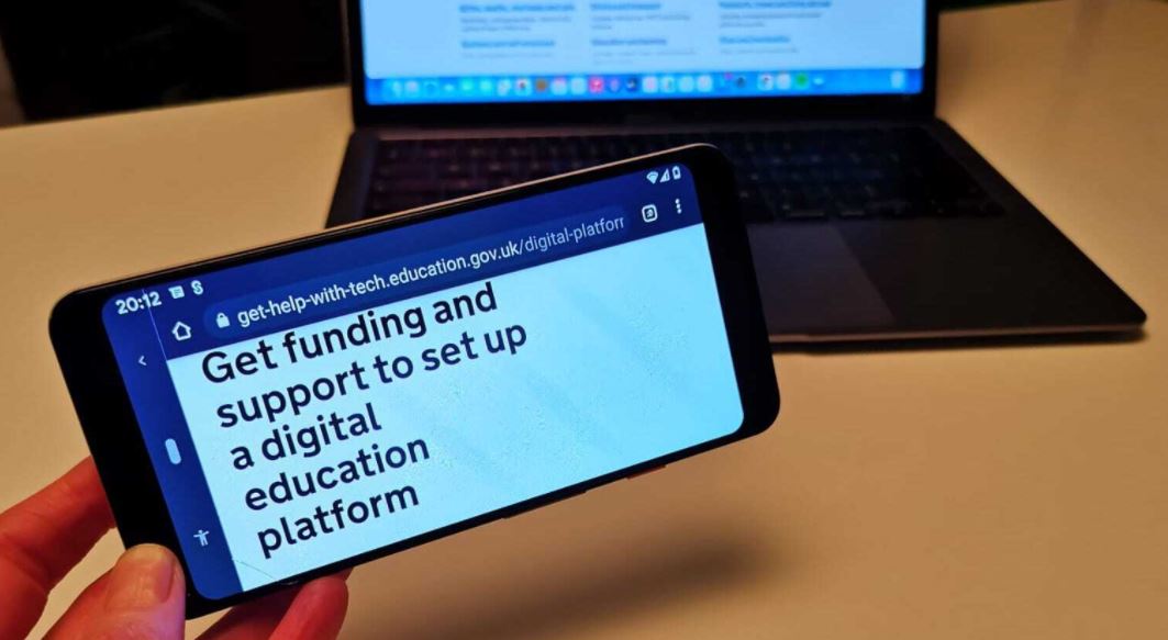 GOV.UK web page on a mobile phone screen in front of a laptop. Mobile phone reads &#39;Get funding and support to set up a digital education platform&#39;.